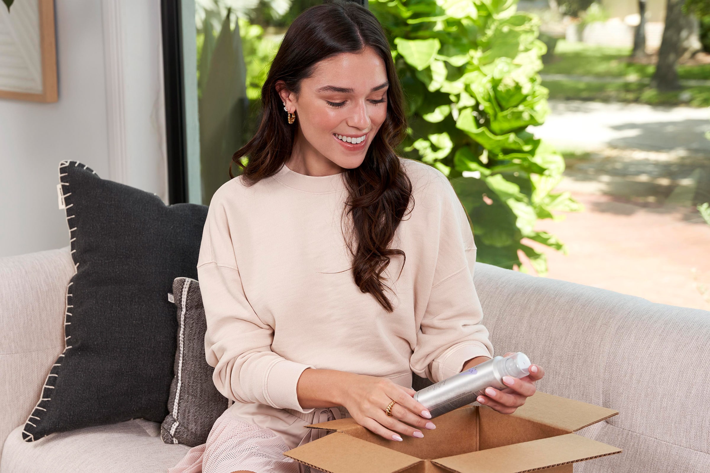 A woman with dark brown hair sitting on a couch, smiling while holding a MONAT Product bottle, set in a cozy, natural-lit living room