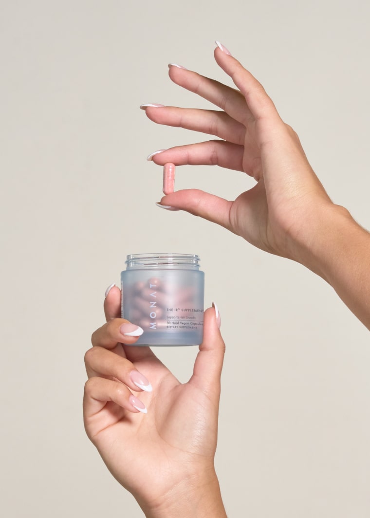 Hands holding pink supplement capsule above open MONAT IR HAIR SUPPLEMENT