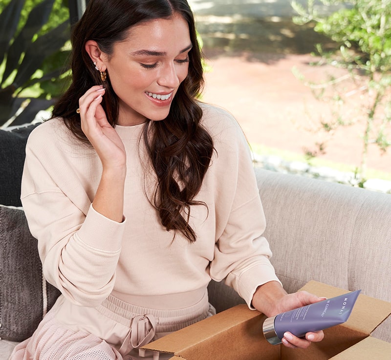 A woman with dark brown hair sitting on a couch, smiling while holding a MONAT Product bottle, set in a cozy, natural-lit living room