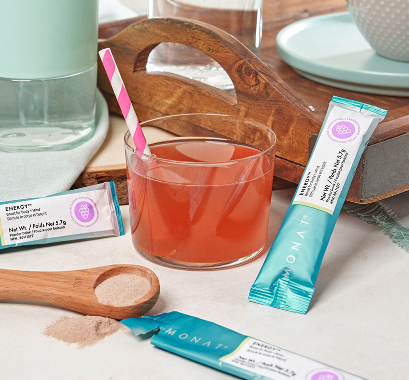 A glass of water, mixed in with MONAT Energy™ Natural Berry and a pink and white straw on a flat surface next 
         to three MONAT Energy™ Natural Berry stick packs, a wooden spoon, a wooden tray, a teal plate, a glass of water and a white bowl. 