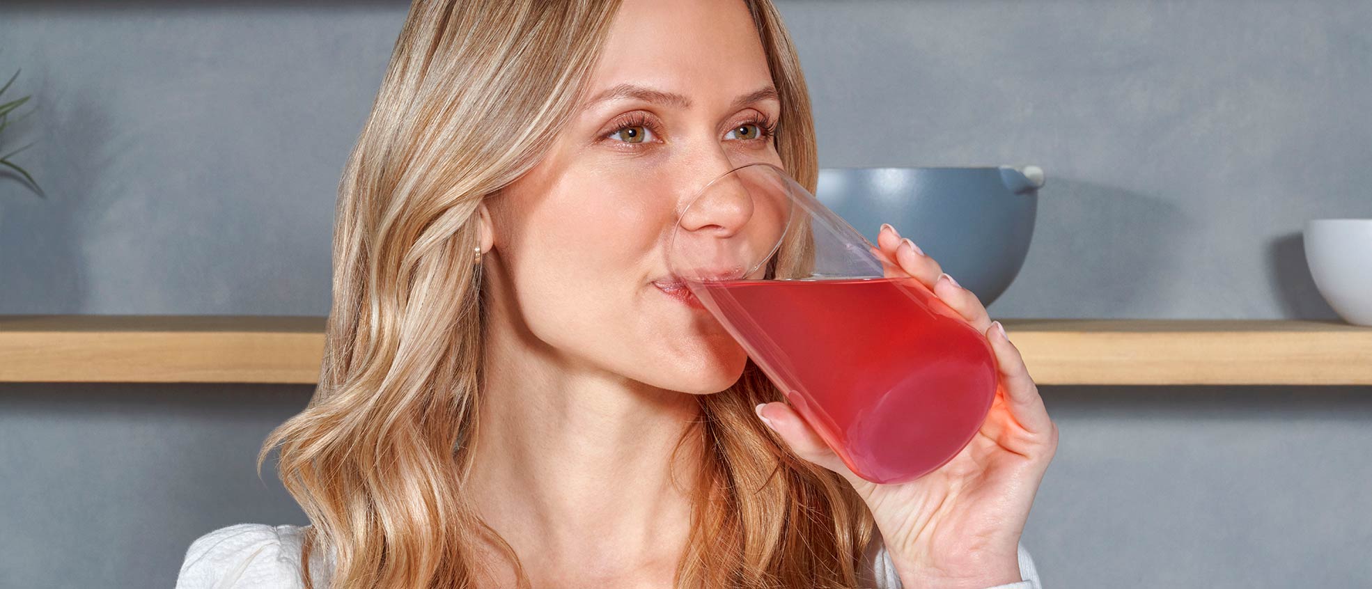 Blonde woman standing in a kitchen and drinking MONAT Energy™ out of a glass cup.