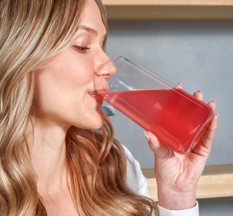 A blonde woman drinking a  glass of water, mixed in with MONAT Energy™ Natural Berry, overlayed on an image of green coffee beans. 