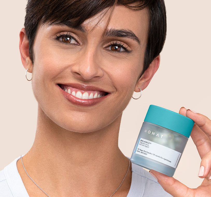 A brunette female smiling while holding MONAT Balance, overlapping a close up photo of pieces of ginger.