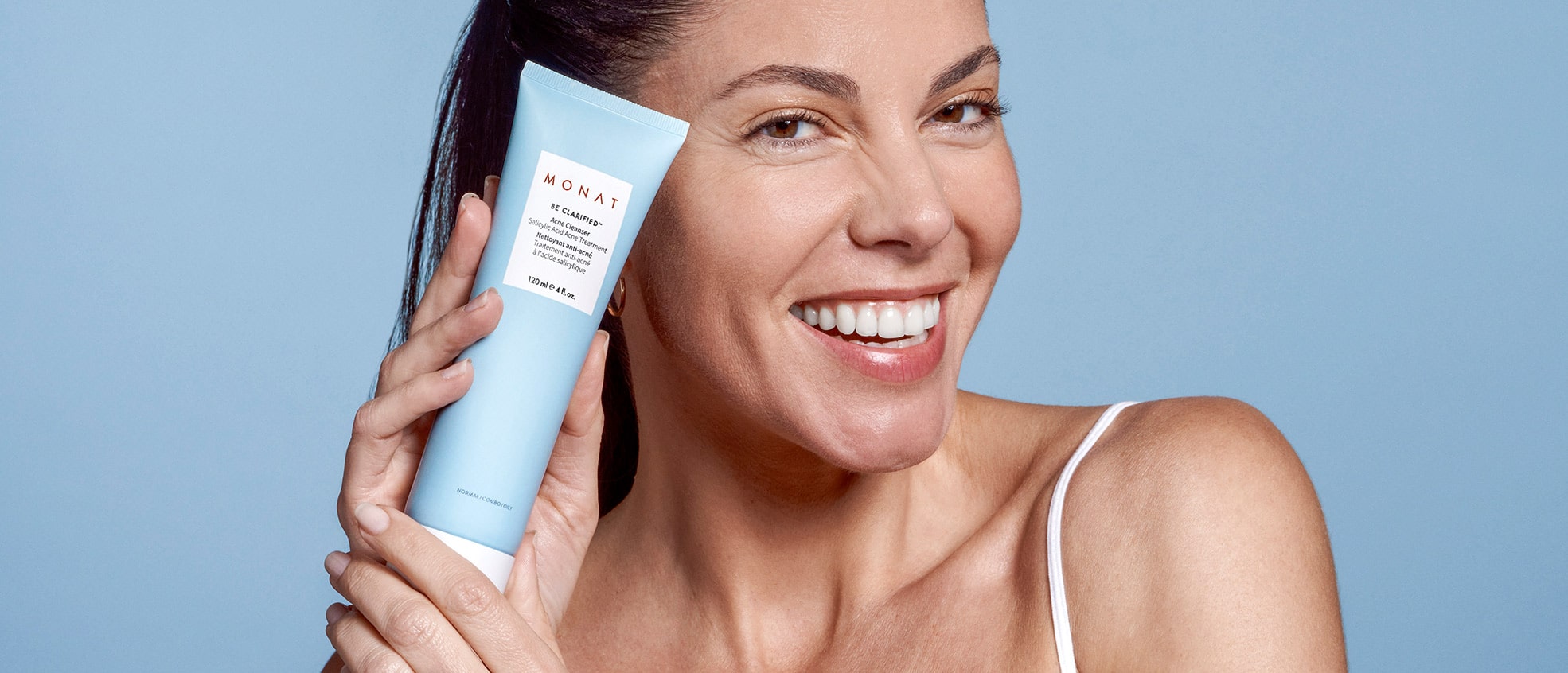 A female smiling while holding BE CLARIFIED™ Acne Cleanser next to her cheek.