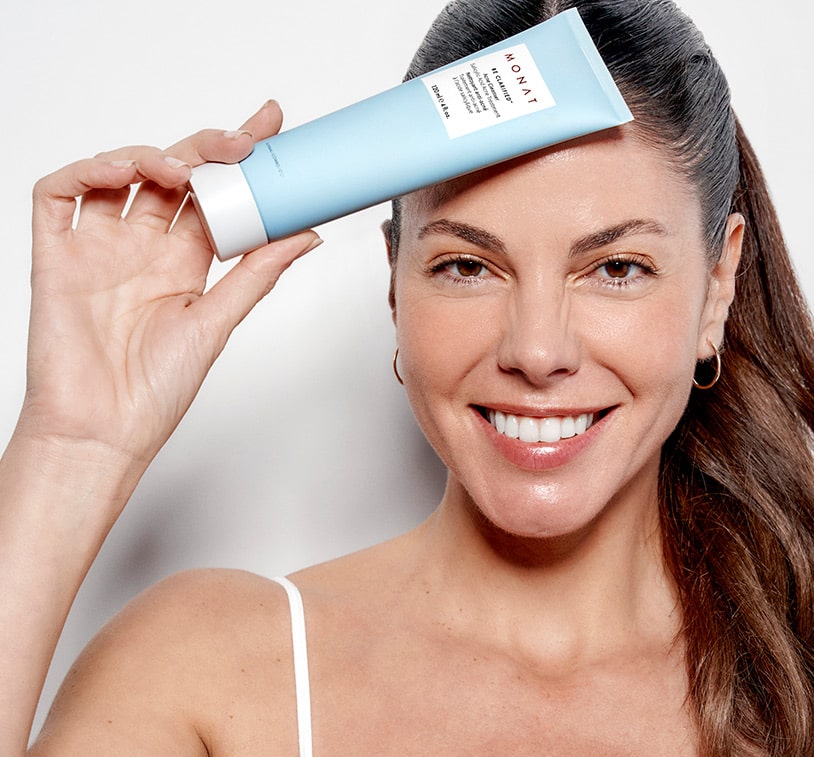 A brunette female holding BE CLARIFIED™ Acne Cleanser infront of her forehead, while overlaying a texture shot of the cleanser. 