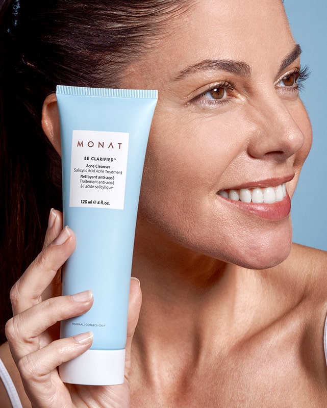A brunette female smiling while holding BE CLARIFIED™ Acne Cleanser next to her cheek. 