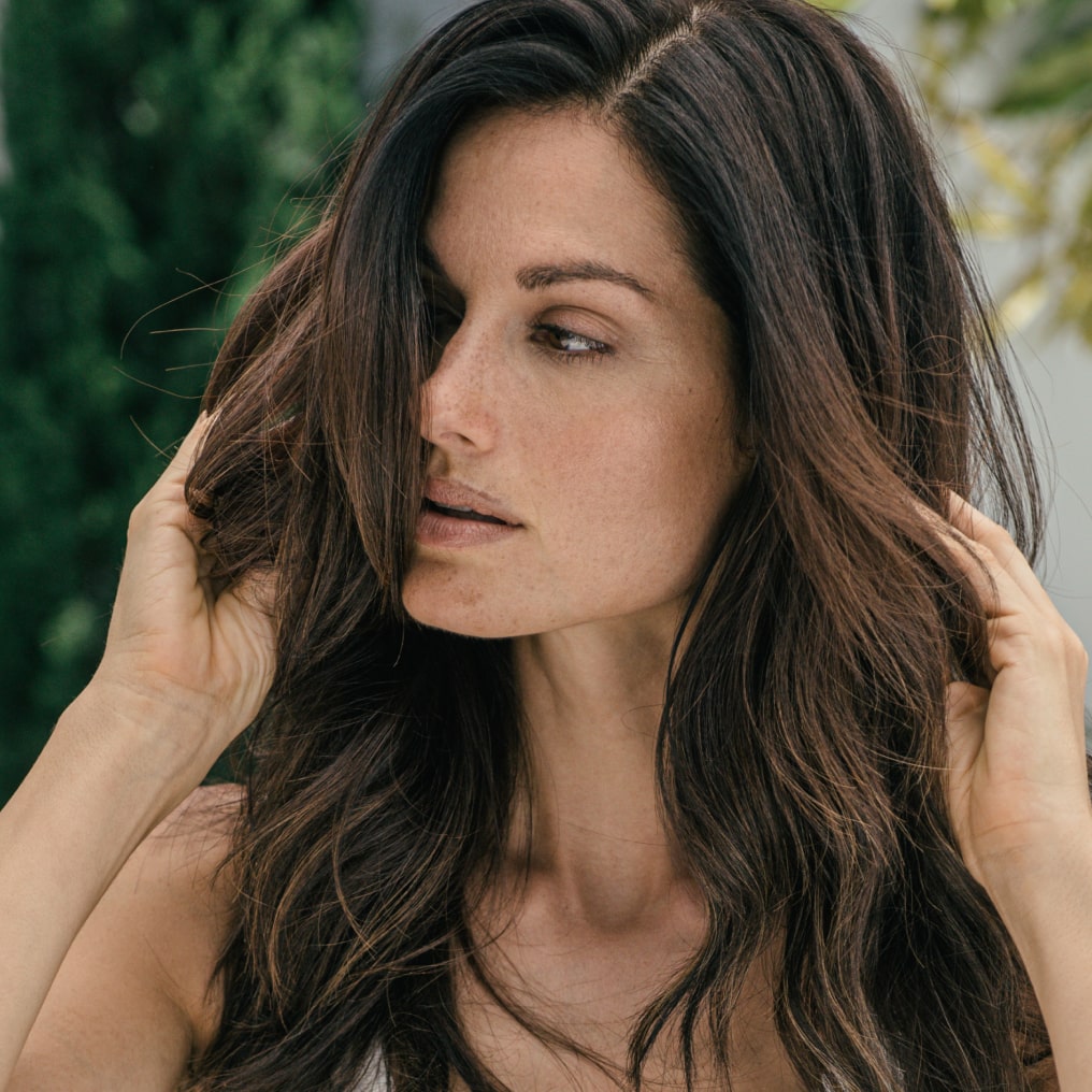 A close-up of a woman with long, wavy brown hair, gently touching her hair, smiling softly.