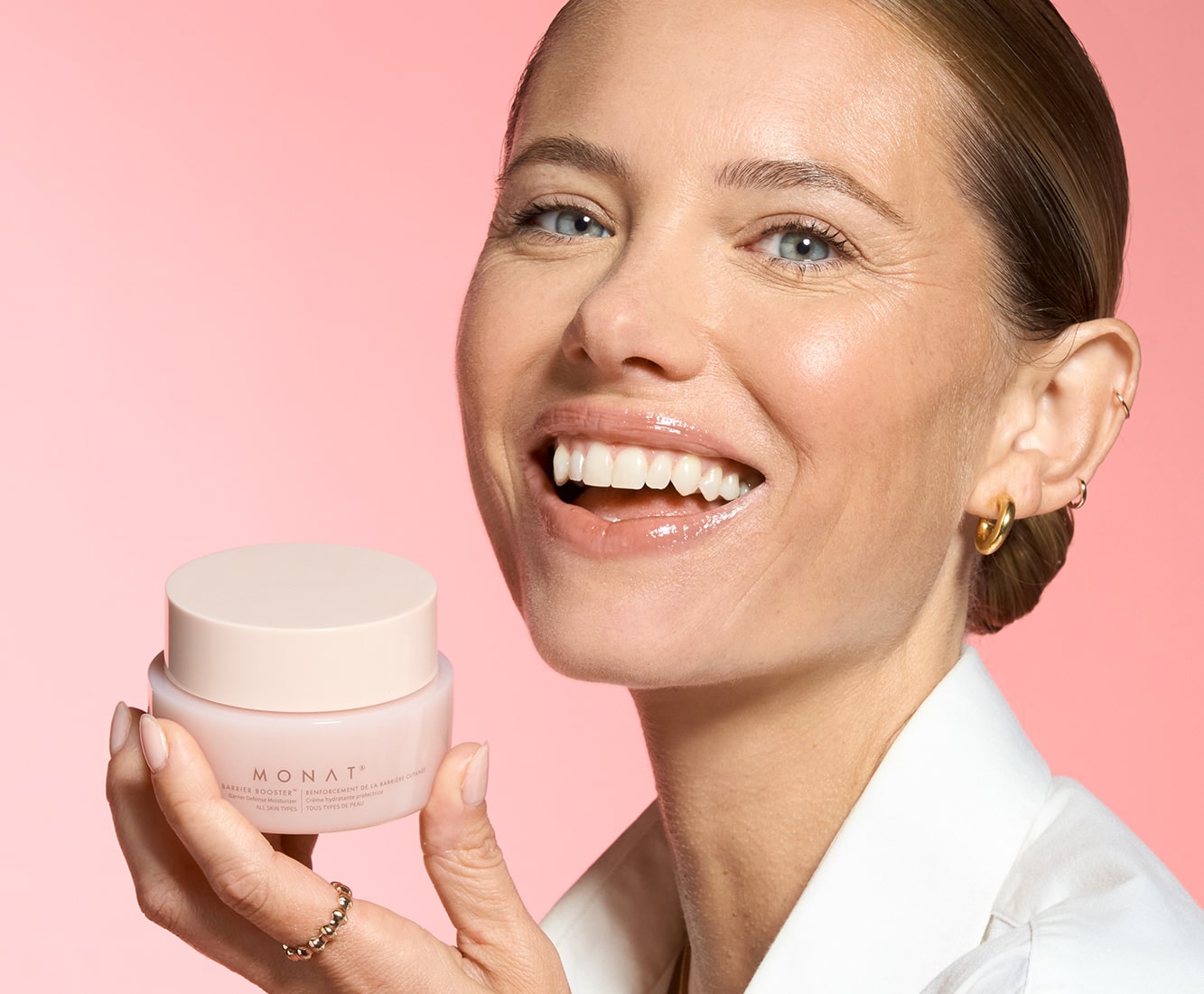 Smiling woman holding MONAT Barrier Booster™ Barrier Defense Moisturizer jar near her cheek.