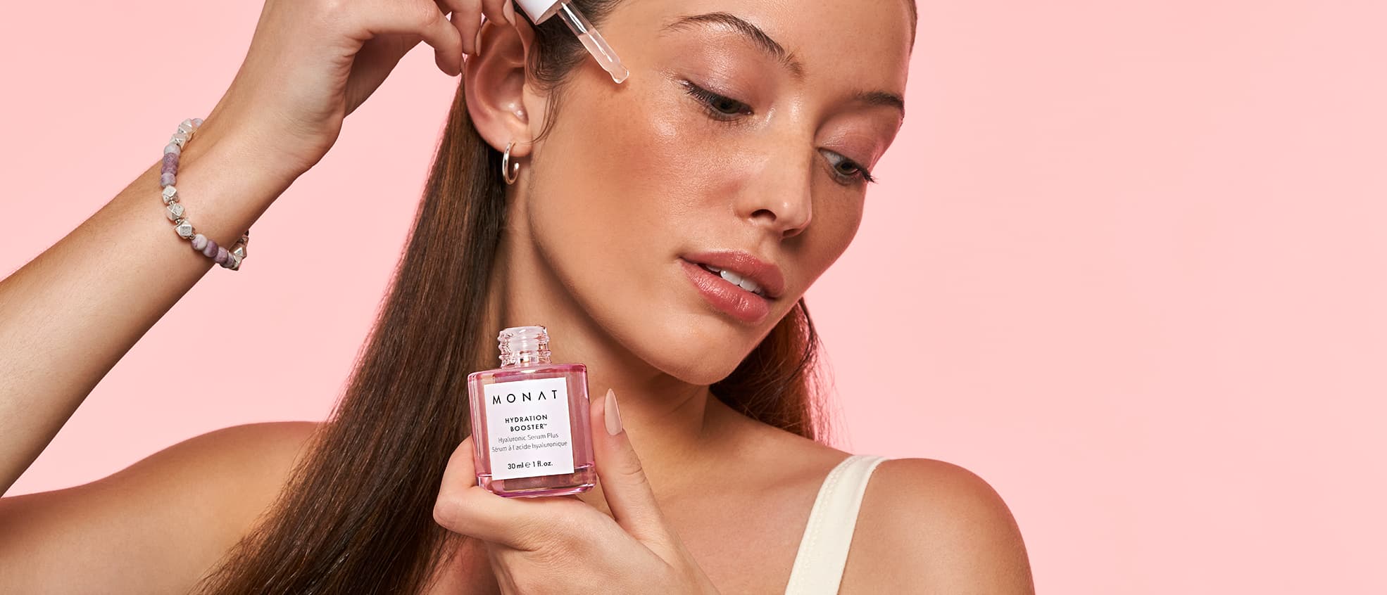 Woman smiling while looking to her left side. High contrast image.  Holding MONAT's Hydration Booster on her left hand while applying 
      the eyedropper on her cheeks.