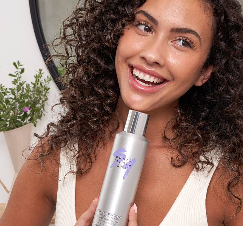 Brunette female smiling and holding MONAT STUDIO ONE™ Flex Control Hairspray.