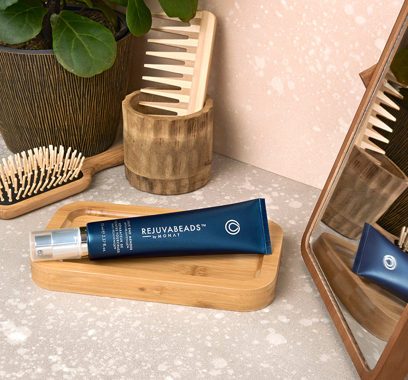 REJUVABEADS® by MONAT laying on a wooden tray sitting on a counter next to a mirror, plant, a wooden comb and wooden brush