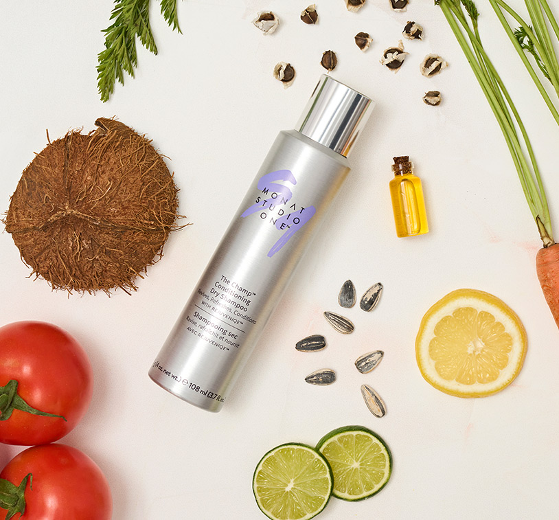 MONAT STUDIO ONE™ The Champ Conditioning Dry Shampoo laying on a flat surface, along with ingredients such as lemon and lime slices, tomatoes, coconut pieces and sunflower seeds.