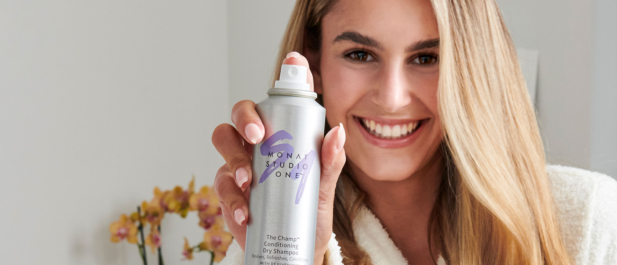 Blonde female smiling while holding MONAT STUDIO ONE™ The Champ Conditioning Dry Shampoo.