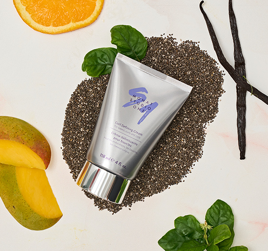 MONAT STUDIO ONE™ Curl Defining Cream on a flat surface with different ingredients such as mangoes, chia seeds, orange slices, and greenery.