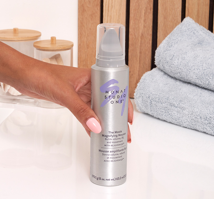 A hand grabbing MONAT STUDIO ONE™ The Moxie™ Magnifying Mousse on a white counter, next to gray bath towels and two glass containers.