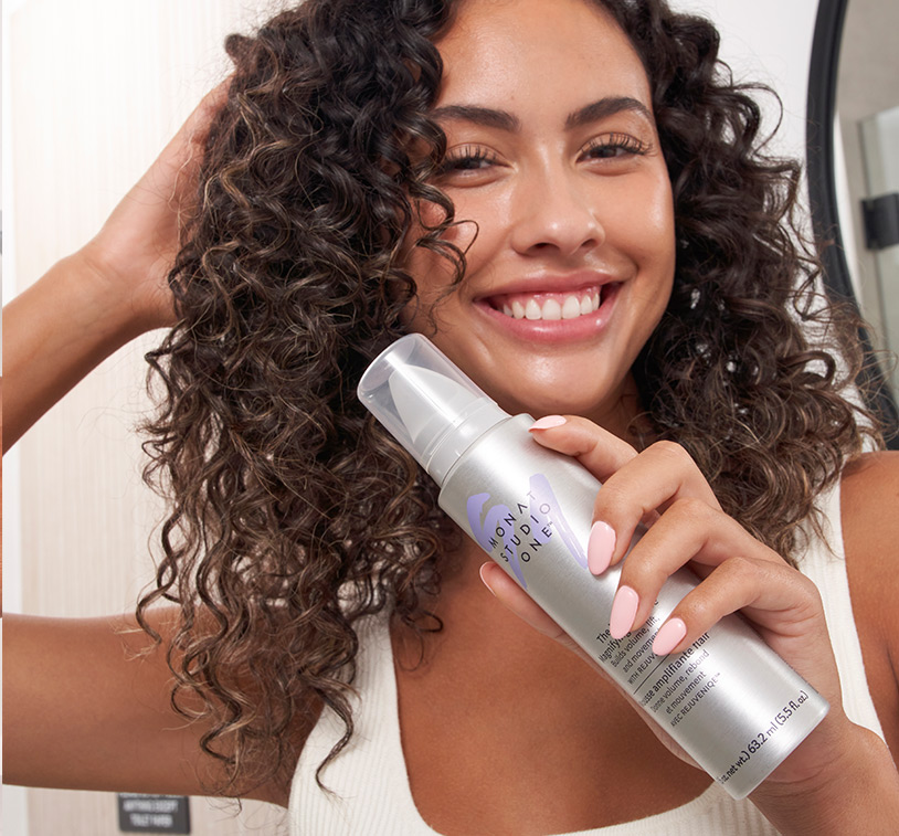 Brunette female with curls holding MONAT STUDIO ONE™ The Moxie Magnifying Mousse.