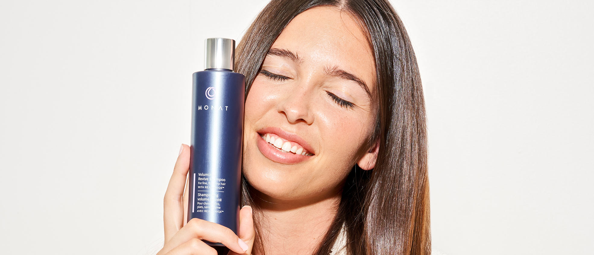 Brunette female smiling and holding the Volumizing Revive Shampoo against her face.