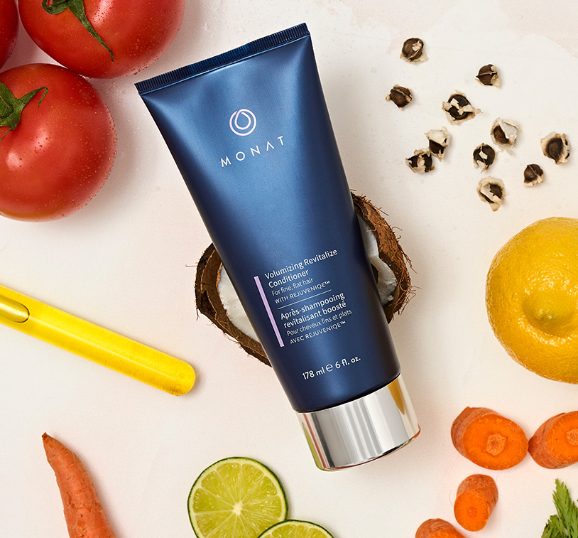 Volumizing Revitalize Conditioner laying on a flat surface, along with ingredients such as lemon slices, coconut pieces, tomatoes, carrots and oil.