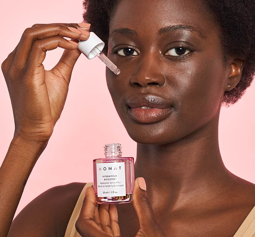 An african-american woman looking directly at the camera while applying the serum with the eyedropper. Left hand 
      showcases MONAT's Hydration Booster.