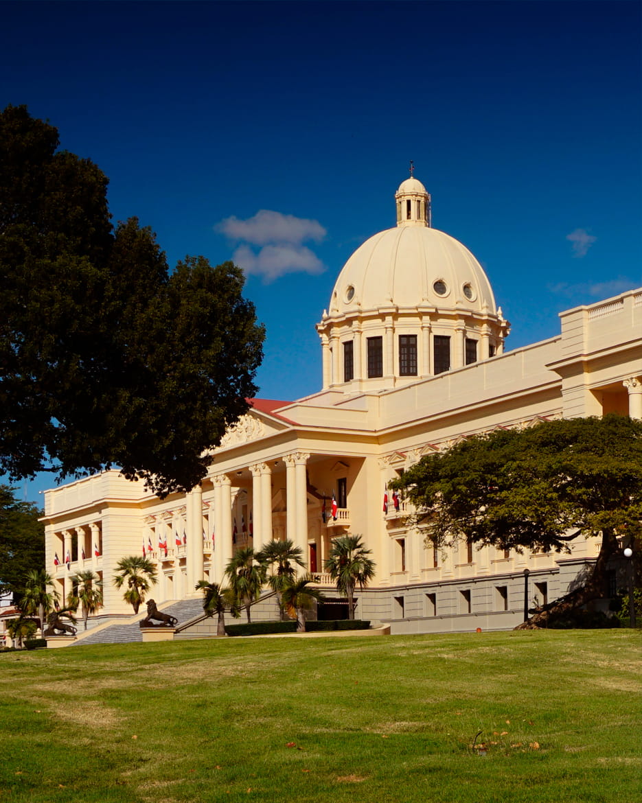 Neoclassical government building with a large white dome and columns in Santo Domingo, Dominican Republic, surrounded by palm trees and a green lawn.