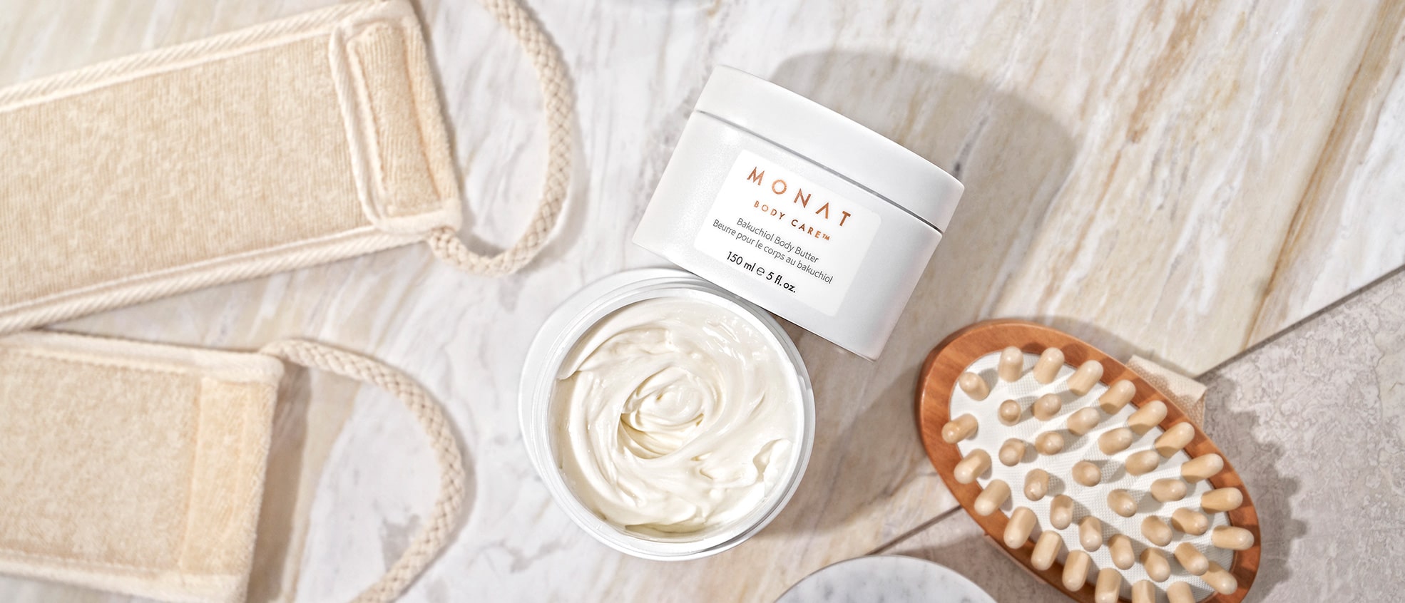 Two jars of a MONAT BODY CARE™ Bakuchiol Body Butter, one of this open showing its content, surrounded of a hair brush and a band of towel.