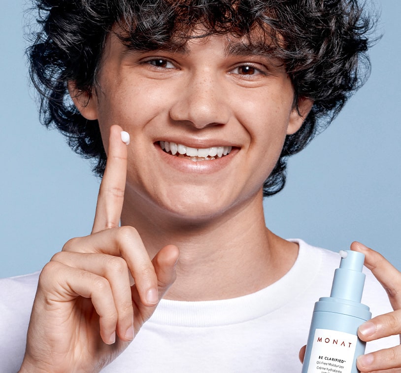 A male smiling while holding an opened BE CLARIFIED™ Acne-Clearing Moisturizer, with some moisturizer on his finger. 