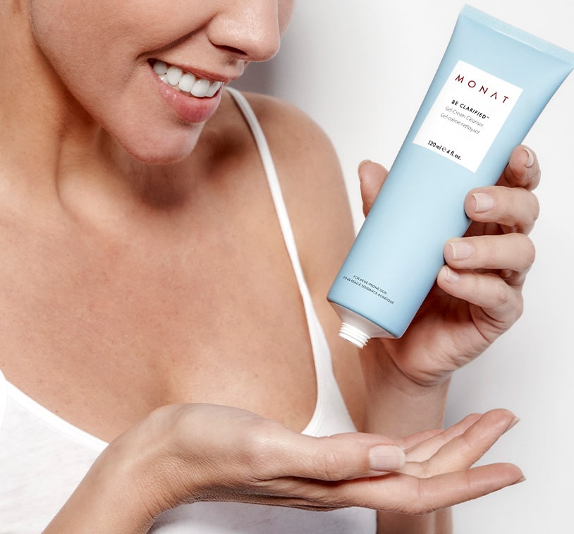 A female wearing a white tank top holding an opened bottle of BE CLARIFIED™ Acne Gel-Cream Cleanser. 