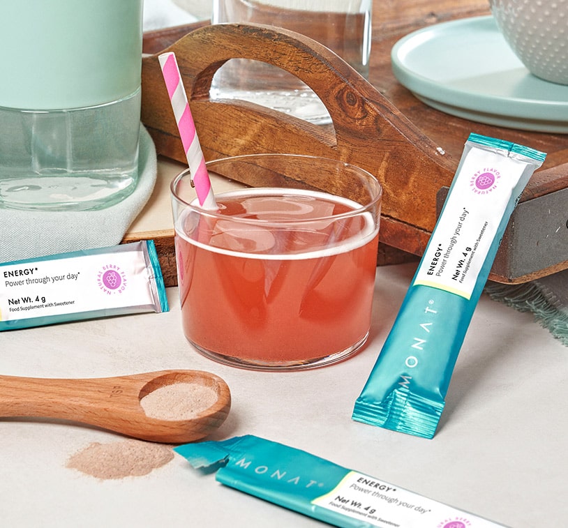 A glass of water, mixed in with MONAT Energy™ Natural Berry and a pink and white straw on a flat surface next 
            to three MONAT Energy™ Natural Berry stick packs, a wooden spoon, a wooden tray, a teal plate, a glass of water and a white bowl. 