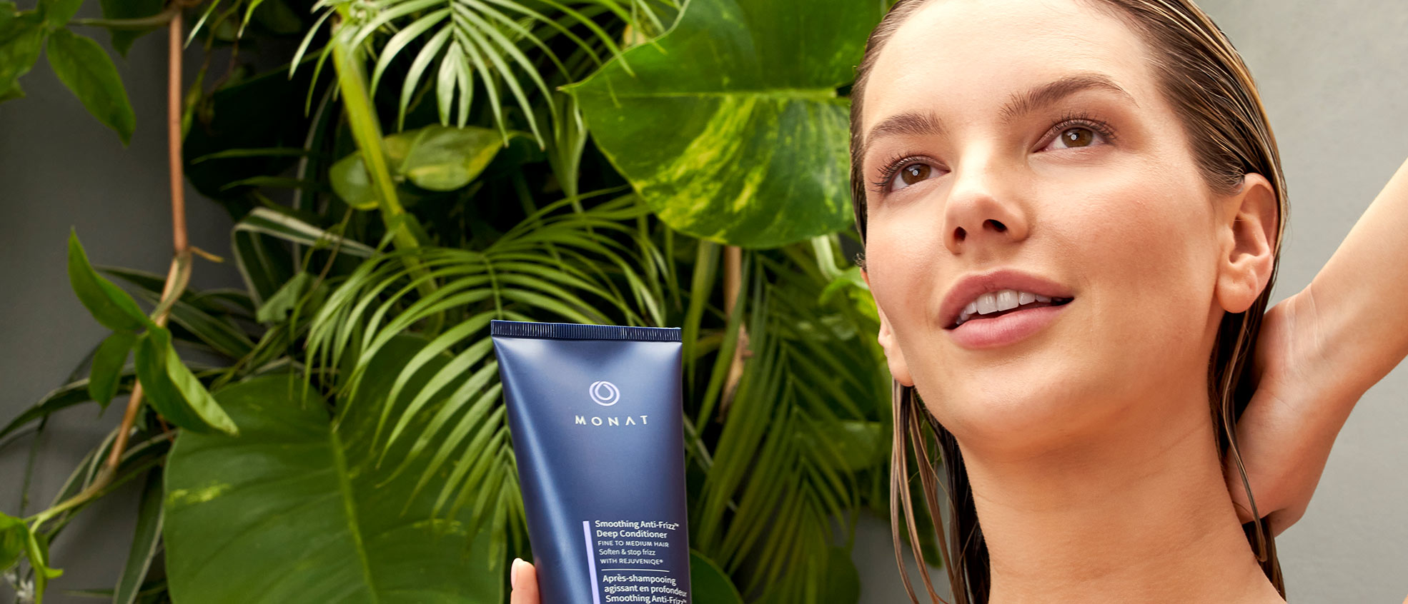 A blonde female with damp hair, standing near greenery and holding Smoothing Anti-Frizz™ Deep Conditioner.