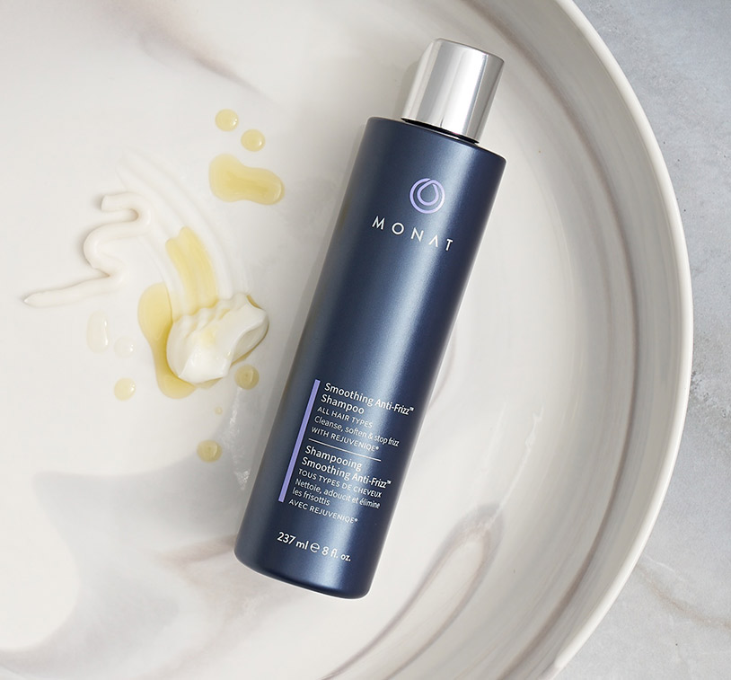 Smoothing Anti-Frizz™ Shampoo laying on a ceramic plate, next to drops of REJUVENIQE® and Smoothing Anti-Frizz™ Shampoo.