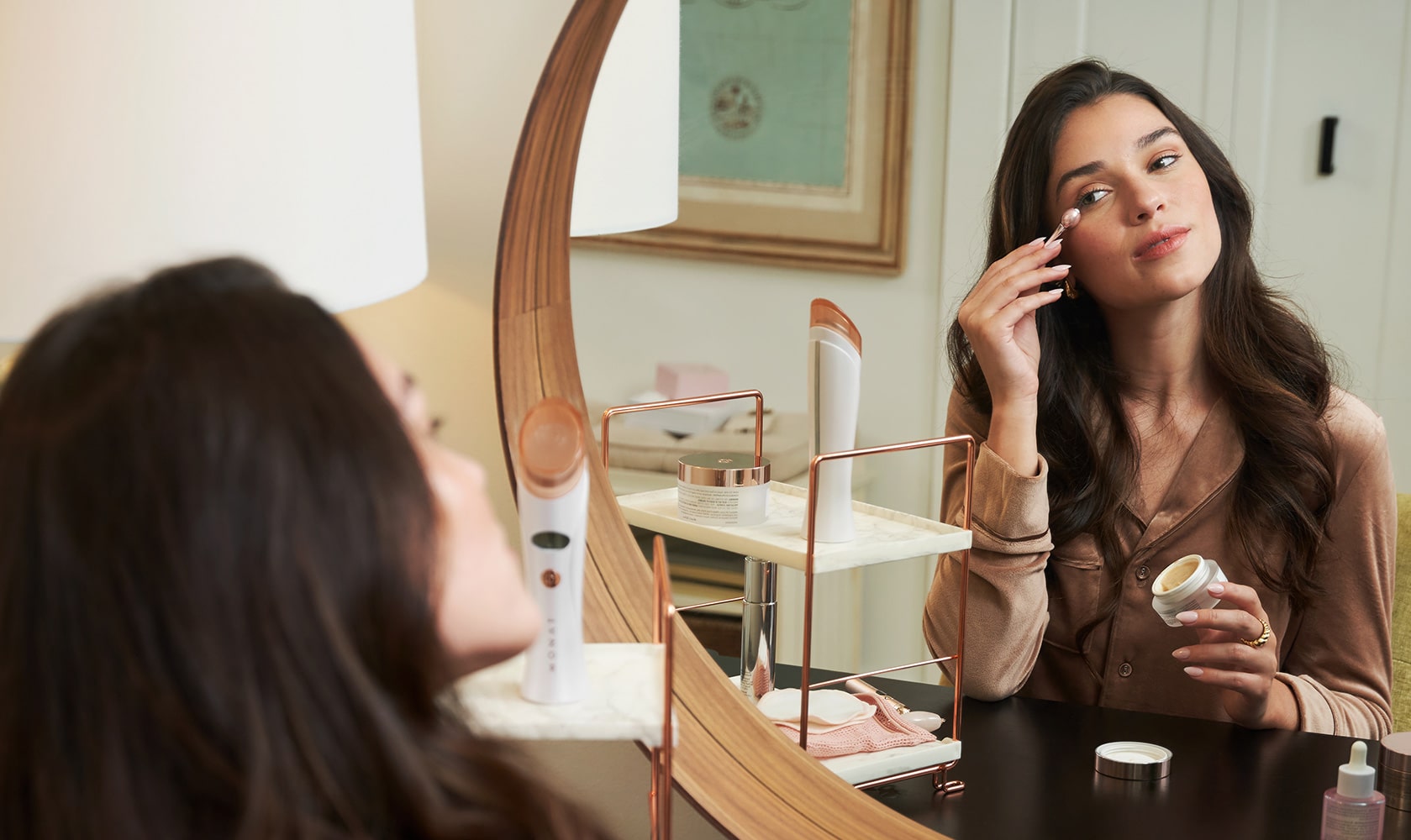 A dark-haired woman in a bathroom, smiling while reaching for a Monat product on a stylish shelf filled with other beauty products.