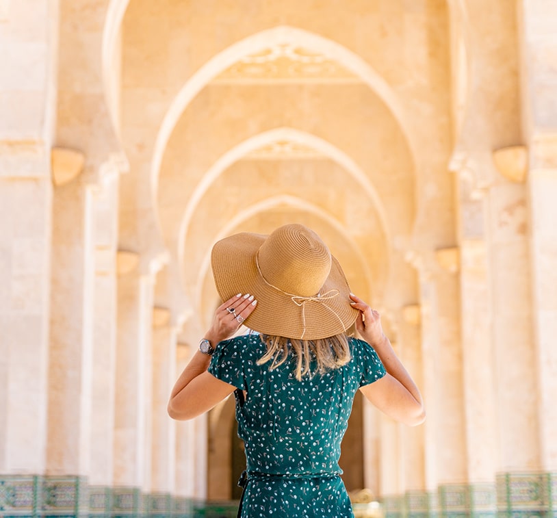 A woman seen from behind, wearing a green dress and holding a large hat in her hands, admires the arched architecture of a hallway with Moroccan-style design.