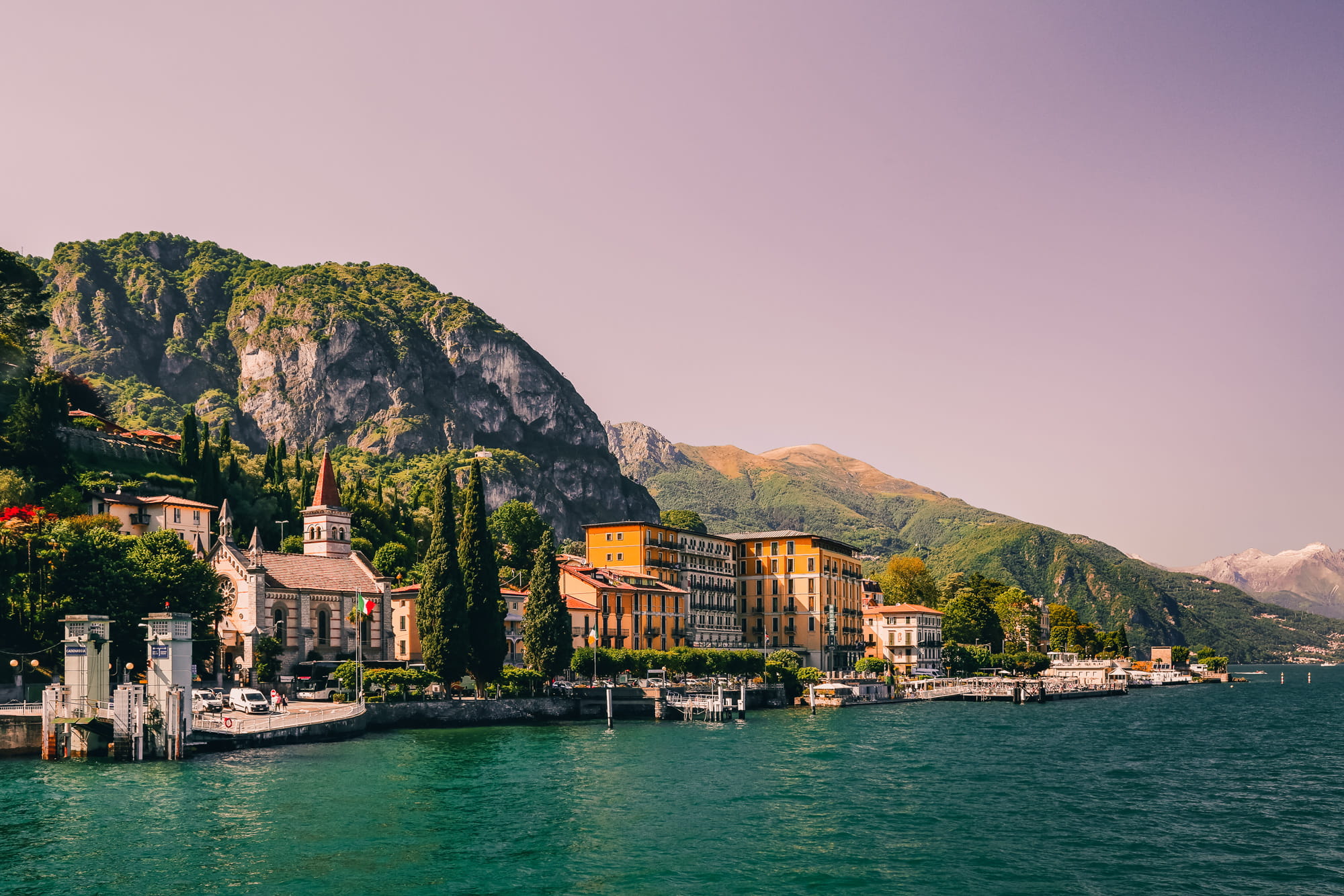 Scenic lakeside town with colorful buildings, a church with a bell tower, and tall cypress trees set against dramatic green mountains under a clear sky.