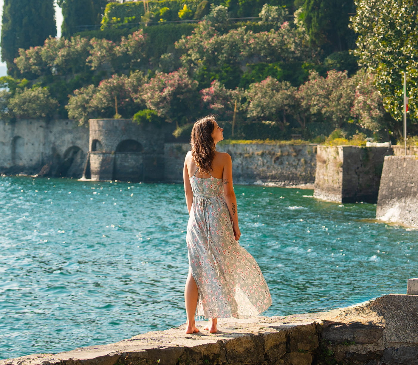 Woman in a light floral dress standing barefoot on a stone edge, looking out over a calm turquoise lake with a historic stone bridge and lush greenery in the background.