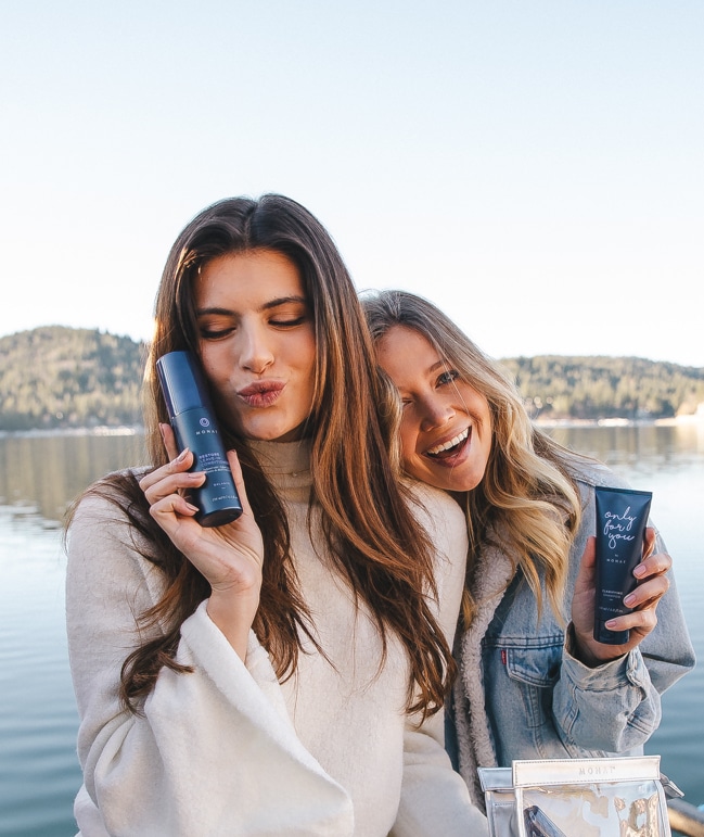Picture of two smiling women standing by a lake and holding MONAT products in their hands.