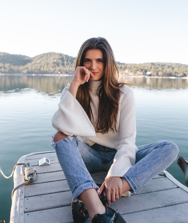 Image of a smiling woman in a white sweater and jeans sitting on pier by a lake