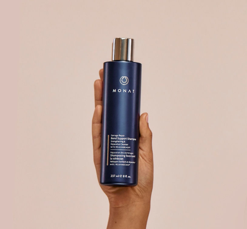 MONAT's Damage Repair Shampoo being upheld by a woman's hand, showcasing the product's aesthetics. Subject is placed in front of a peach color background.
