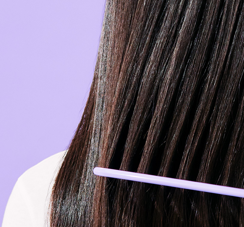 Closeup of a woman’s brunette hair being combed. Depicts smooth results. Subject poses in front of a solid, 
      lavender color background.