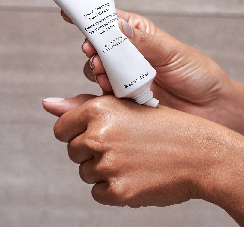 A closeup of a woman applying MONAT's Silky & Soothing Hand Cream onto her hands. This image reveals the cream's white 
      texture.