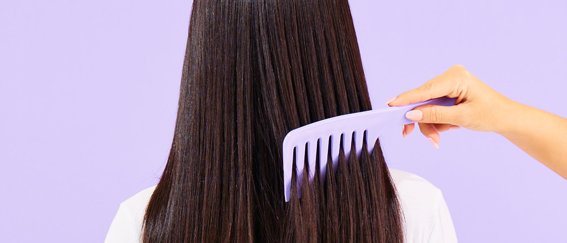 Image shows a woman's back head with brunette hair being combed vertically by another woman's hand. Subject poses in front of a lavender color background.