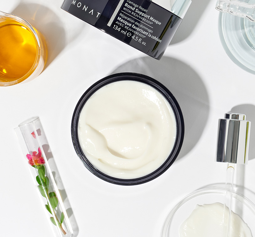 MONAT's Damage Repair Bond Support Masque shown from an aerial perspective with an open lid showing its white texture. Raw ingredients such as oils & herbs along additional glass ornaments are carefully placed around the product.