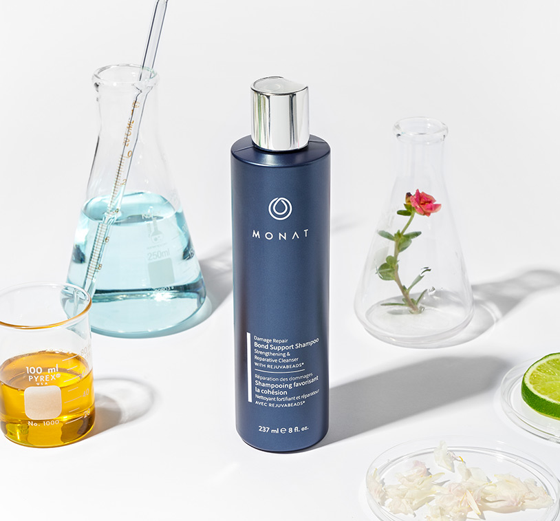 MONAT’s Damage Repair Shampoo stands over a white solid background setting displaying raw ingredients such as oils, citrus & 
         herbs along with additional glass ornaments around it.