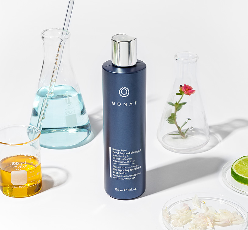 MONAT’s Damage Repair Shampoo stands over a white solid background setting displaying raw ingredients such as oils, citrus & 
				herbs along with additional glass ornaments around it.