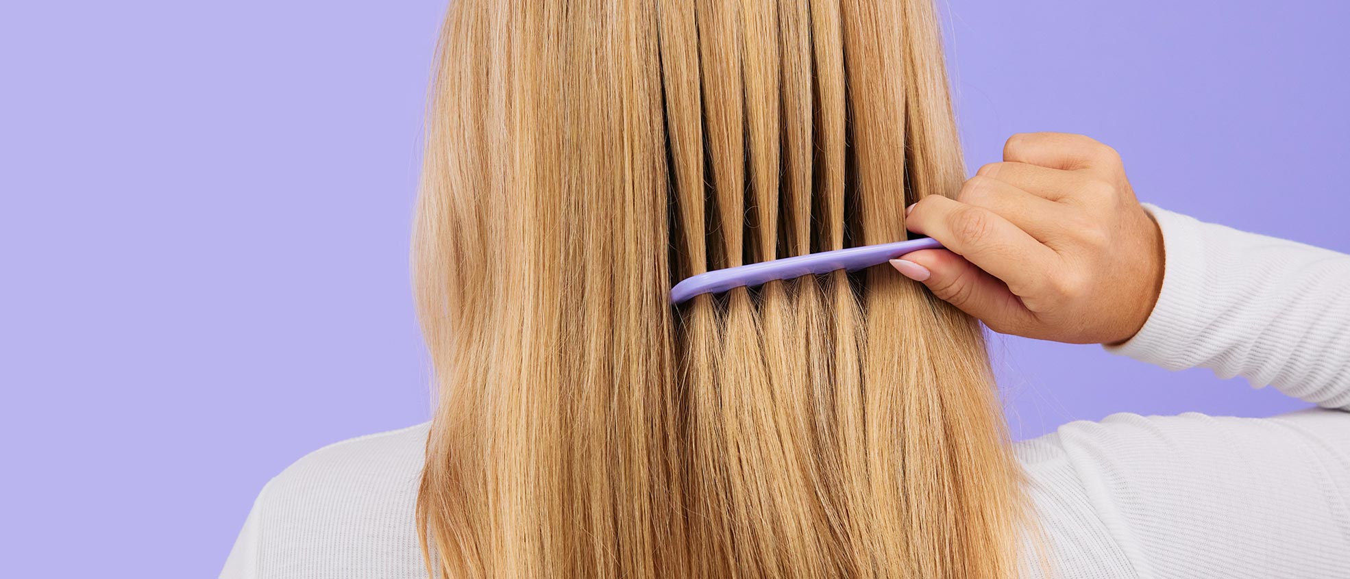 A blond woman’s healthy hair being effortlessly combed with her right hand. Subject poses in front of a lavender color background.