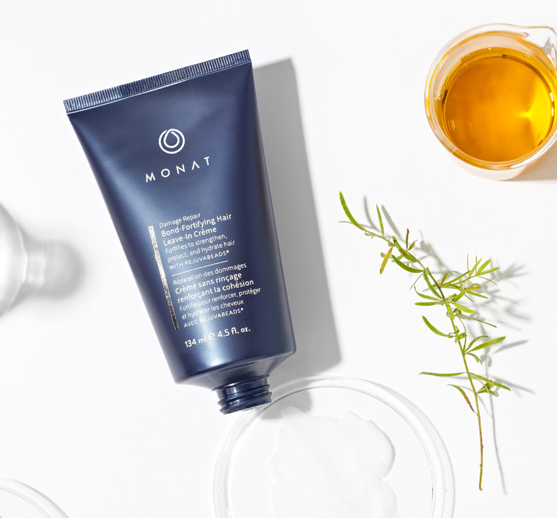 MONAT’s ® Damage Repair Bond-Fortifying Hair Leave-In Crème lays over a solid white background exhibiting 
         its main ingredients such as oils & herbs placed along glass ornaments.