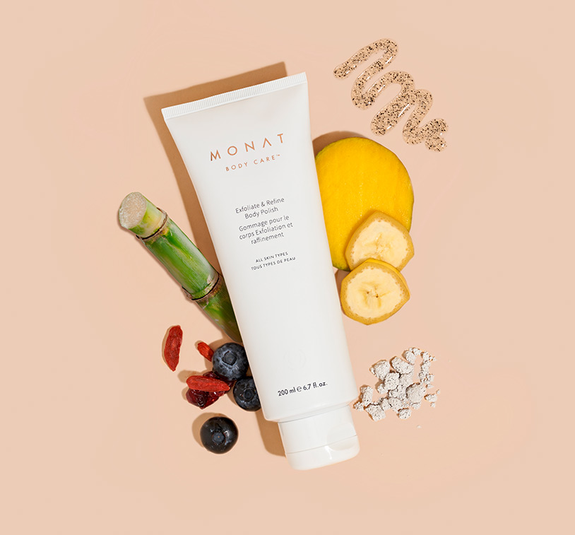 MONAT's Exfoliate & Refine Body Polish lays centered over a solid peach color background showcasing ingredients such as fruits, oils & herbs placed carefully around the main product.