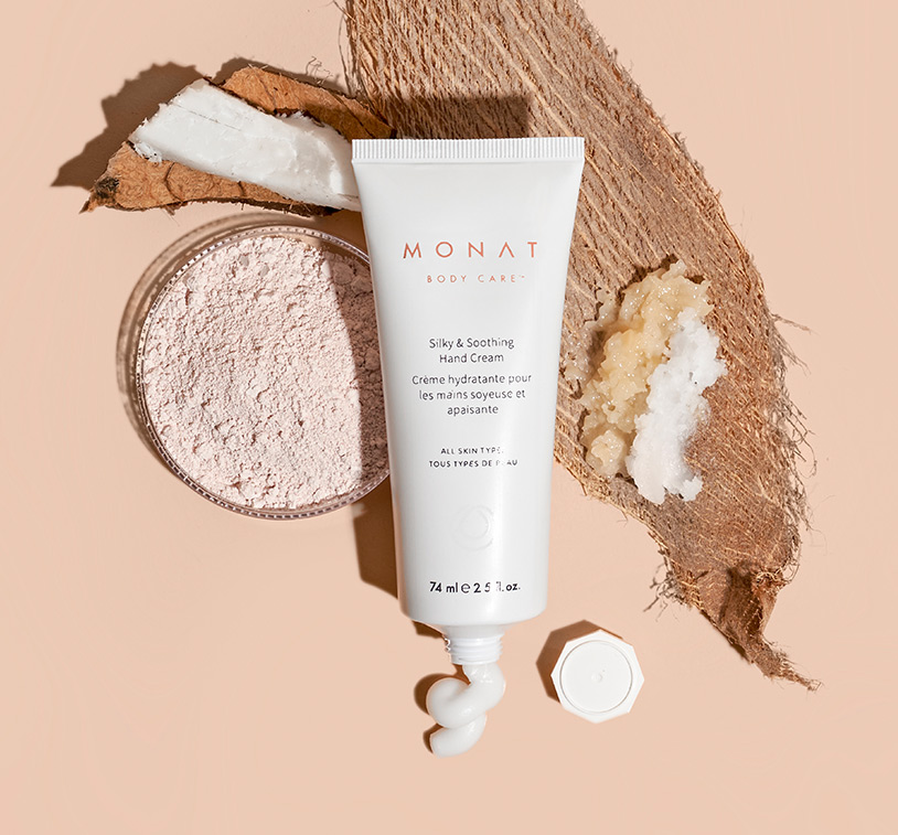 MONAT's Silky & Soothing Hand Cream lays centered over a solid peach color background exhibiting its main ingredients such 
         as coconut & oils ornamenting the main product. 