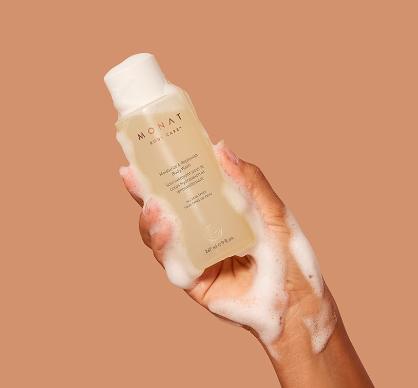 MONAT's Moisturize & Replenish Body Wash being upheld by a woman's hand covered in bath foam, showcasing the 
      product's aesthetics and texture.