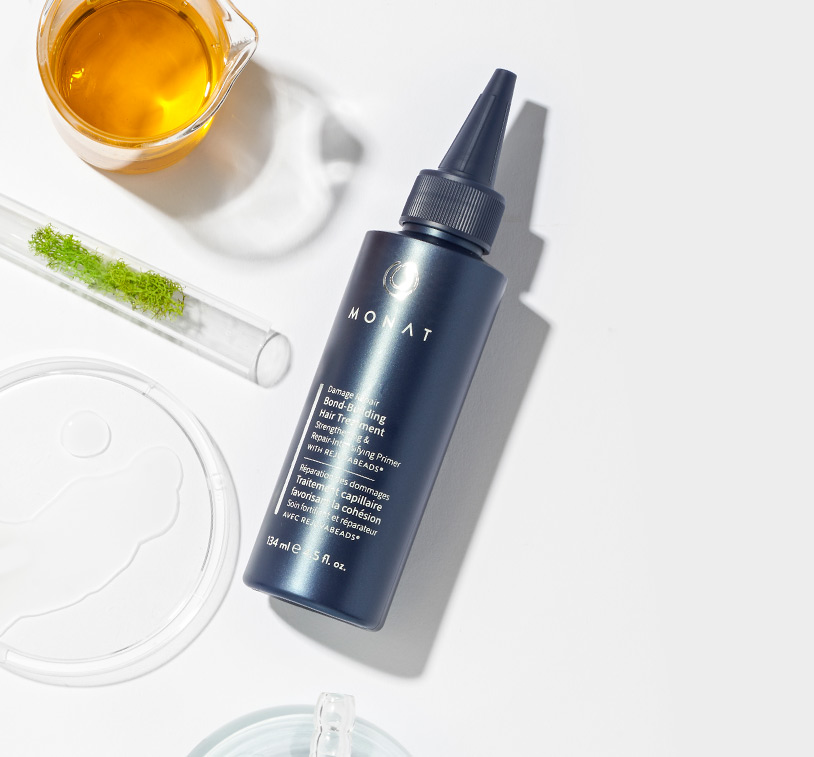 MONAT's Damage Repair Bond-Building Hair Treatment lays over a clean white background setting 
         displaying raw ingredients such as oils & herbs along with additional glass ornaments.