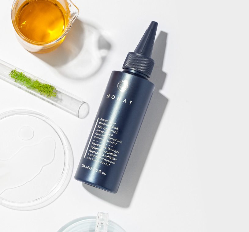 MONAT's Damage Repair Bond-Building Hair Treatment lays over a clean white background setting displaying raw ingredients such as oils & herbs along with additional glass ornaments.